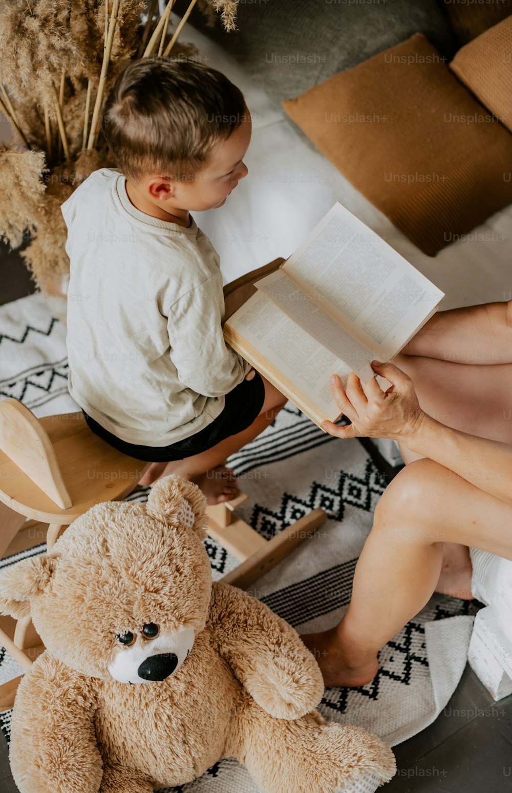 a little boy sitting on a chair reading a book next to a teddy bear