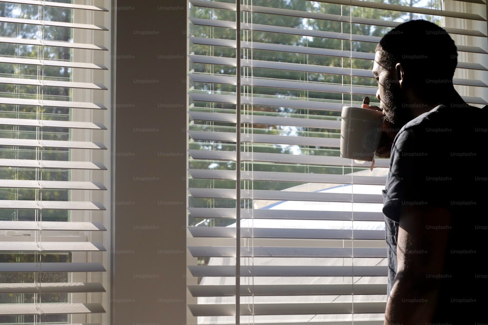 um homem parado em frente a uma janela segurando uma xícara de café