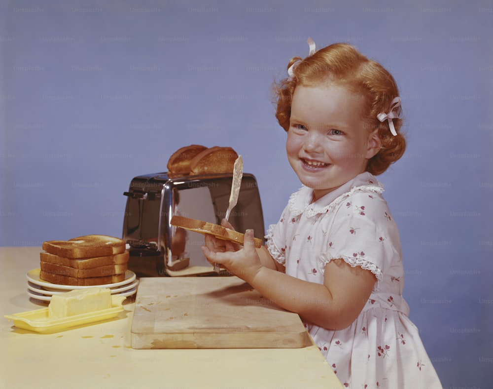 a little girl holding a piece of bread in front of a toaster
