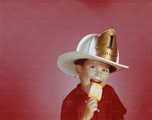 Un giovane ragazzo che indossa un cappello da pompiere e mangia un hot dog