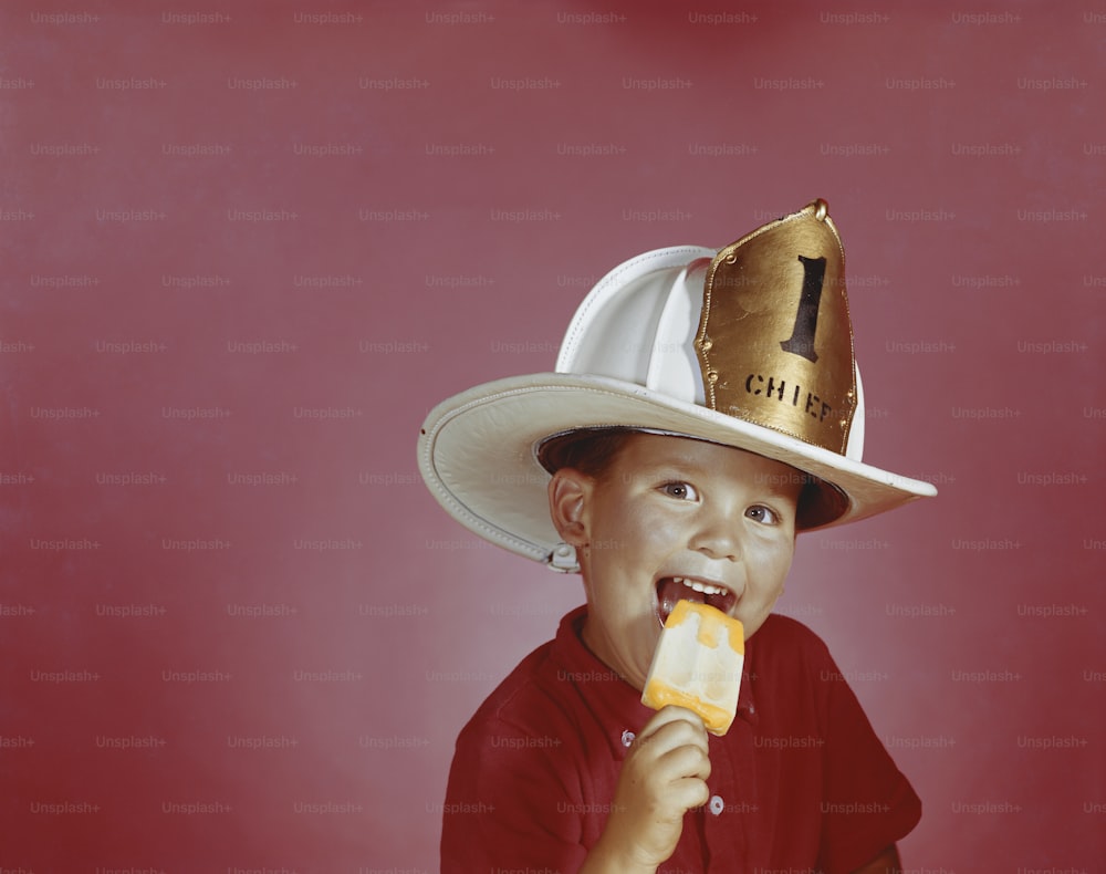 a young boy wearing a fireman's hat and eating a hot dog