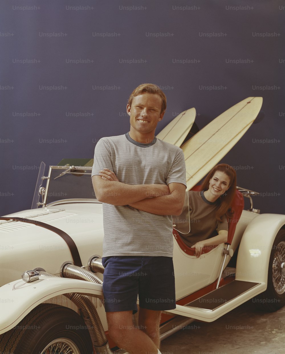 Ein junges Paar mit einem Excalibur Roadster, beladen mit zwei Surfbrettern.