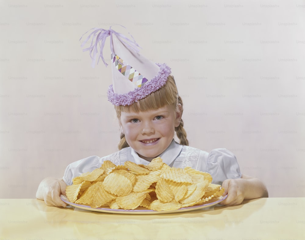 a little girl wearing a party hat holding a plate of chips