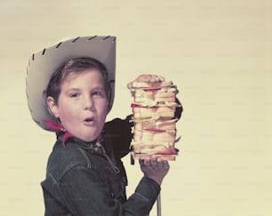 a young boy wearing a cowboy hat holding a stack of sandwiches