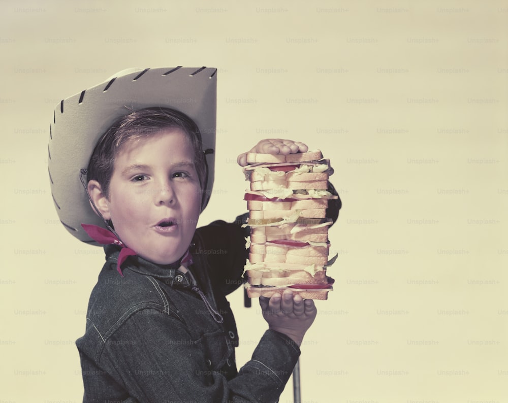 카우보이 모자를 쓴 어린 소년이 샌드위치 더미를 들고 있다