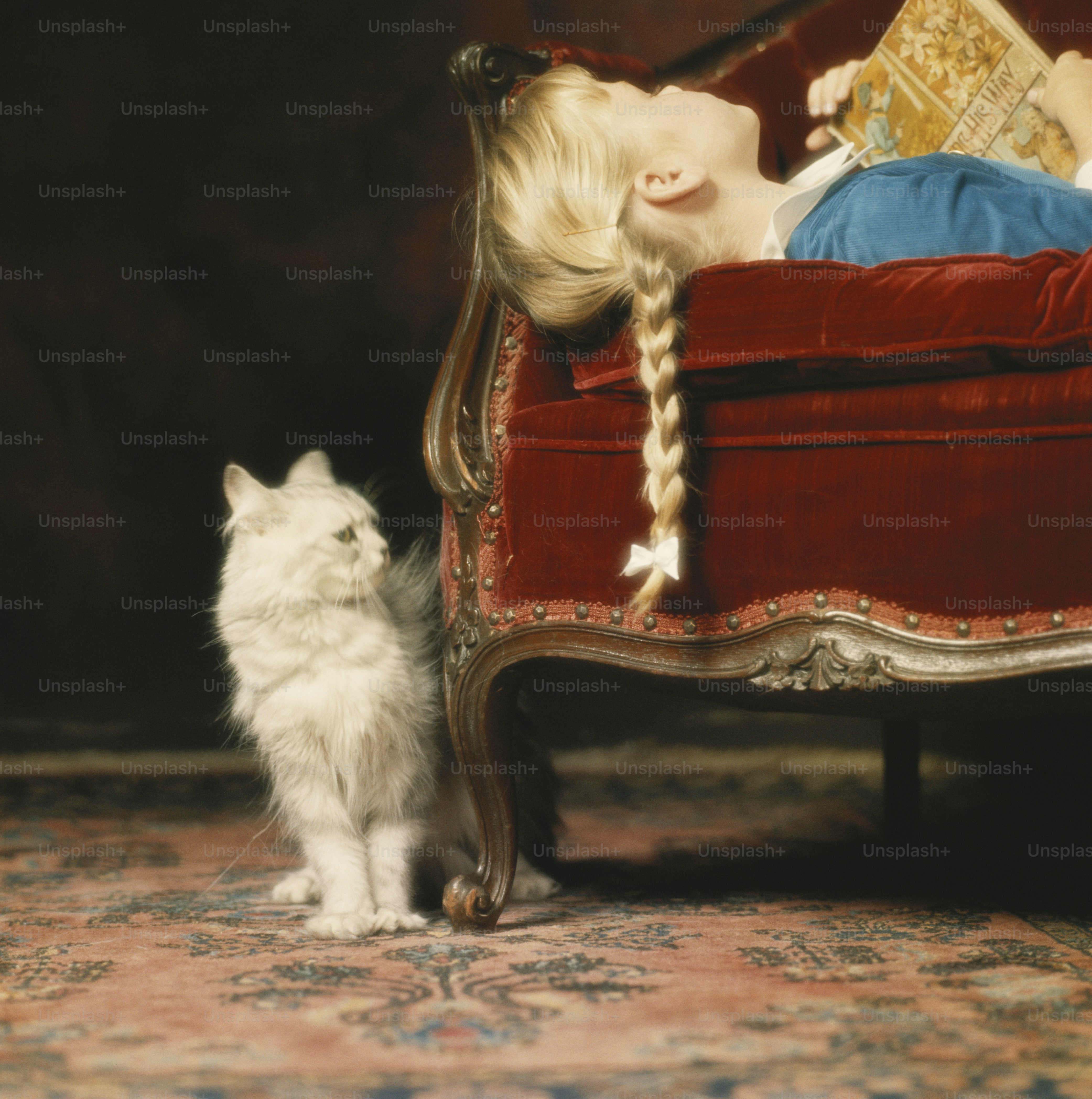 a little girl laying on top of a red couch next to a white cat