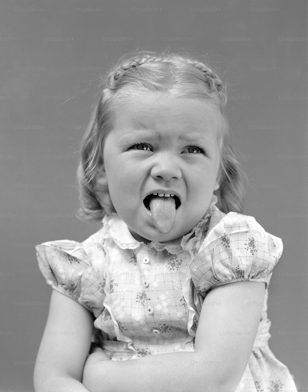 VEREINIGTE STAATEN - Circa 1930er Jahre: Stirnrunzelndes Mädchen mit offenen Armen und herausgestreckter Zunge in einem Kleid mit Baumwollprint und einem weißen Kragen, der in Spitze geschnitten war.