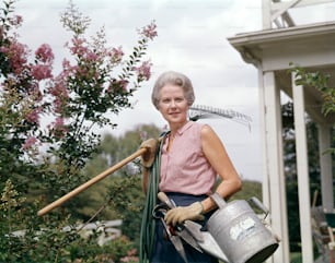 UNITED STATES - Circa 1950s:  Woman Gardener Holding Rake Hose Watering Can Garden Tools Wearing Gloves.