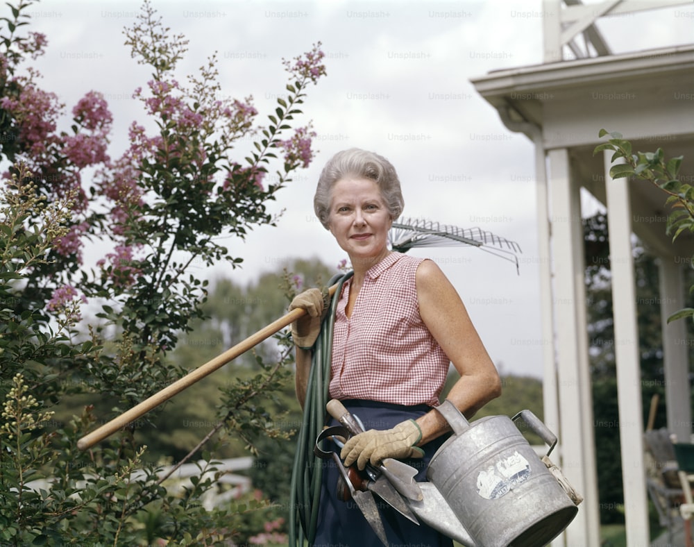 ESTADOS UNIDOS - Por volta de 1950: Mulher jardineira segurando rega de mangueira de ancinho pode ferramentas de jardim usando luvas.