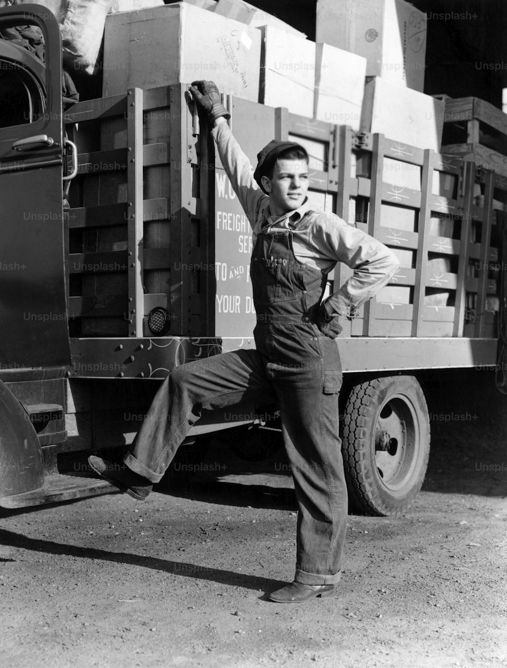 UNITED STATES - Circa 1930s:  Man Truck Driver Dressed In Overalls Gloves Resting Foot On Cab Of Truck And Hand On Truck Bed Loaded With Boxes.