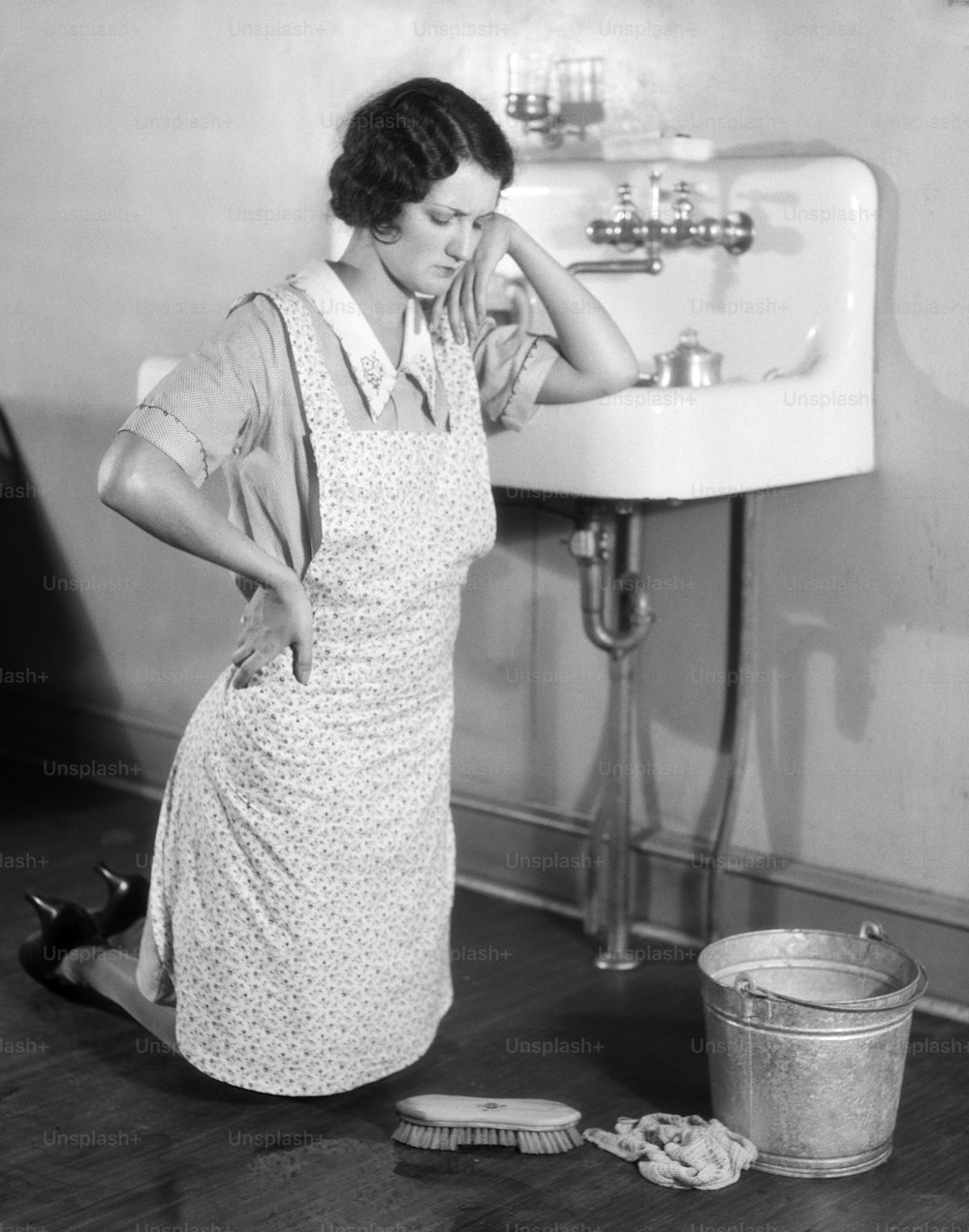 UNITED STATES - CIRCA 1930s: Woman On Her Knees, With Hand On Hip And Elbow On The Sink And A Frown On Her Face. She Is Wearing An Apron Over Her Dress, With Scrub Brush And Pail On The Floor, With Rag.  (Photo by H. Armstrong Roberts/Retrofile/Getty Imag