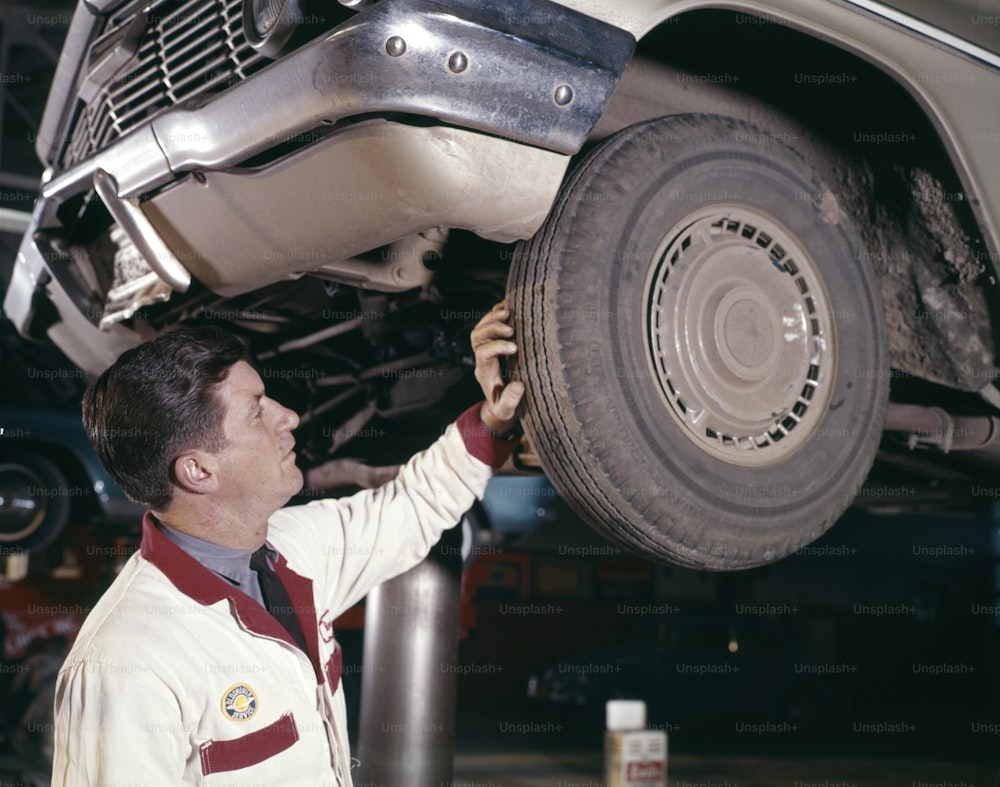 アメリカ合衆国 - 1960年代頃:ガソリンスタンドのリフトラックに車が乗っているときにゴムタイヤのトレッドをチェックするガソリンスタンドの自動車整備士の男性。