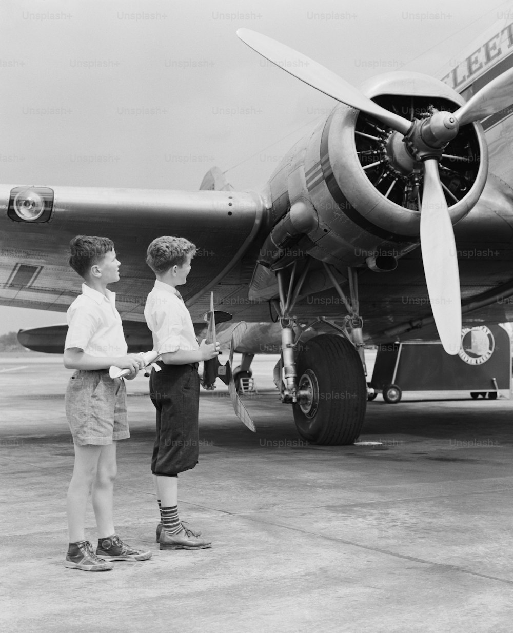 ESTADOS UNIDOS - POR VOLTA DE 1940: Dois meninos de pé ao lado de um avião a hélice, segurando um avião de brinquedo.