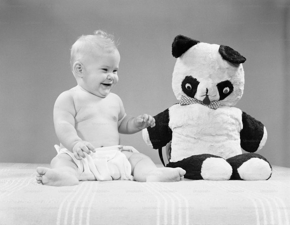 UNITED STATES - CIRCA 1950s:  Baby sitting next to stuffed panda bear toy, smiling and laughing.