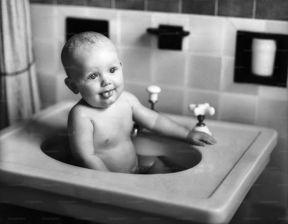UNITED STATES - CIRCA 1950s:  Baby sitting in porcelain sink in bathroom, sticking out tongue.