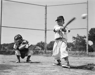 アメリカ合衆国 - 1960年代頃:ユニフォームを着たキャッチャーがしゃがんでボールに手を伸ばし、スイングの途中でボールを打つ少年。