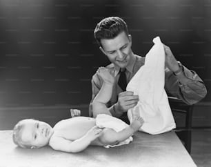 UNITED STATES - CIRCA 1940s:  Young father with baby lying on table, trying to figure out how to put on diaper.
