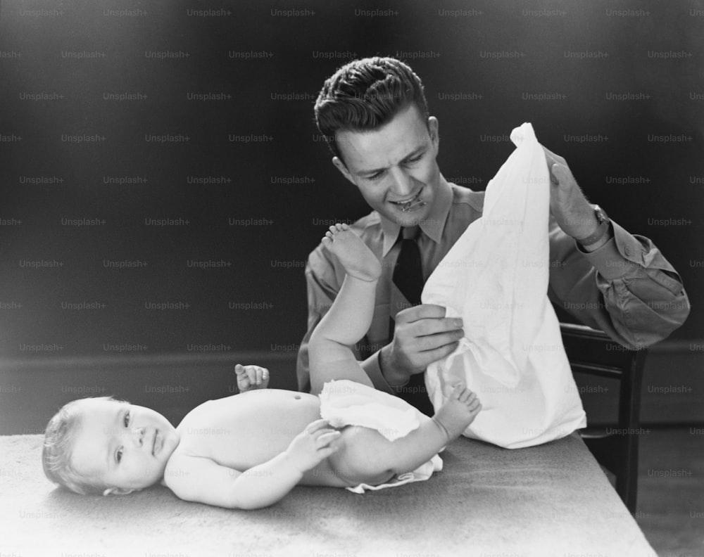 UNITED STATES - CIRCA 1940s:  Young father with baby lying on table, trying to figure out how to put on diaper.
