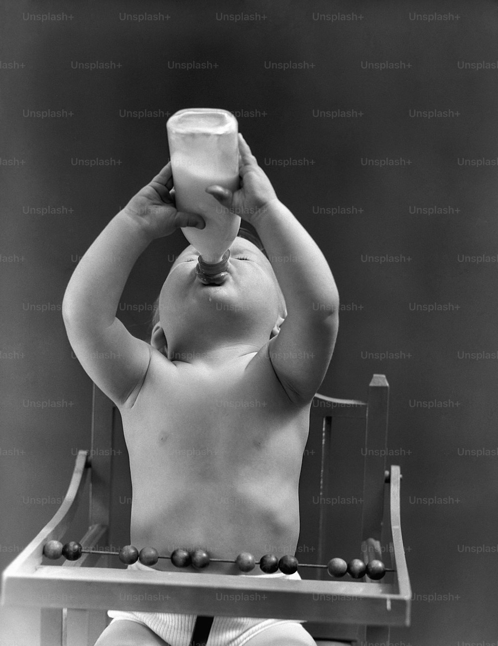 UNITED STATES - CIRCA 1940s:  Baby sitting in high chair, head tilted back, drinking from milk bottle.