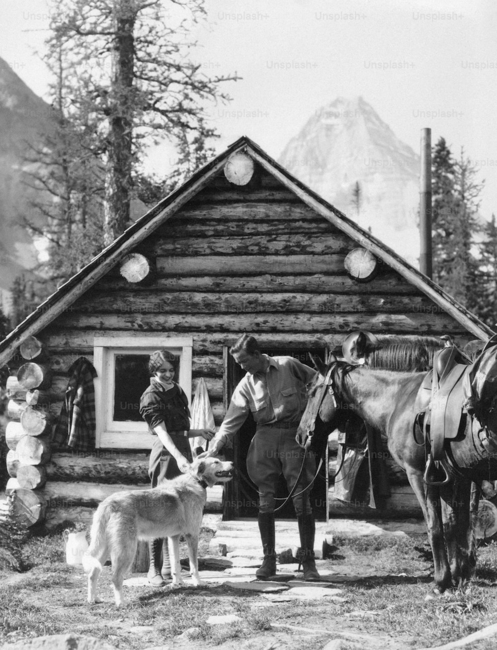 UNITED STATES - CIRCA 1920s:  Couple petting dog in front of log cabin.