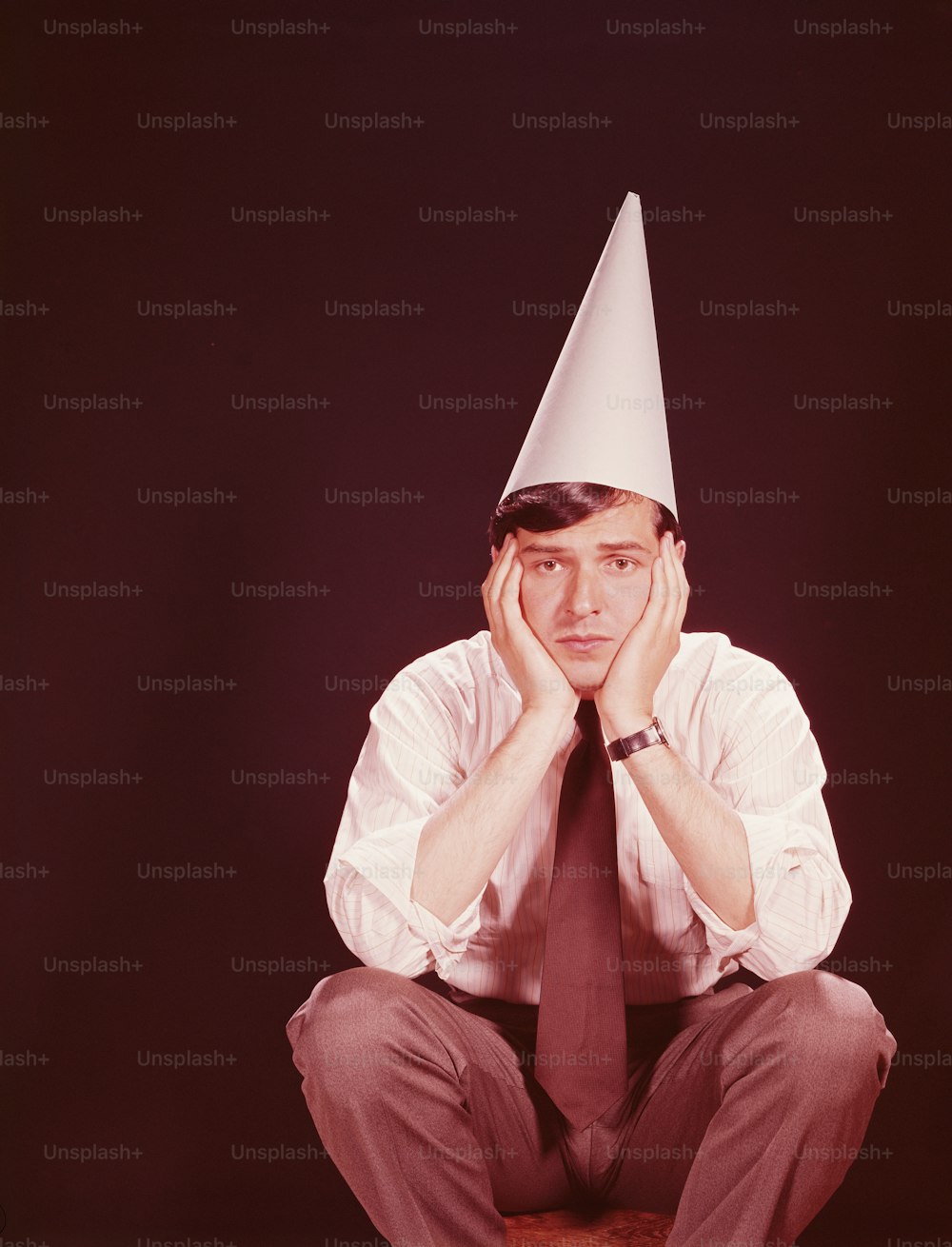 UNITED STATES - CIRCA 1970s:  Man sitting on stool, wearing dunce's hat.