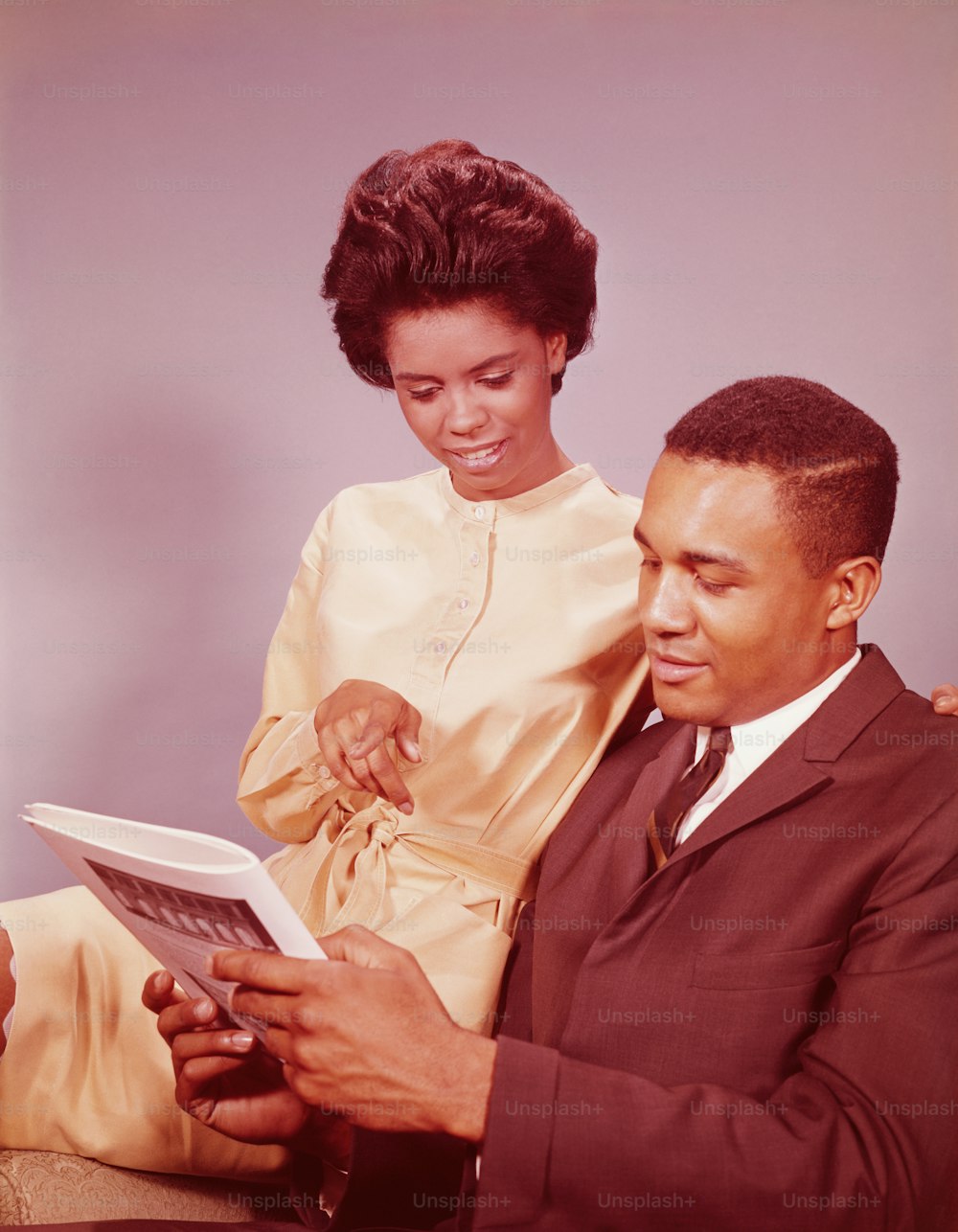 UNITED STATES - CIRCA 1960s:  Couple sitting on chair, woman pointing at magazine article.