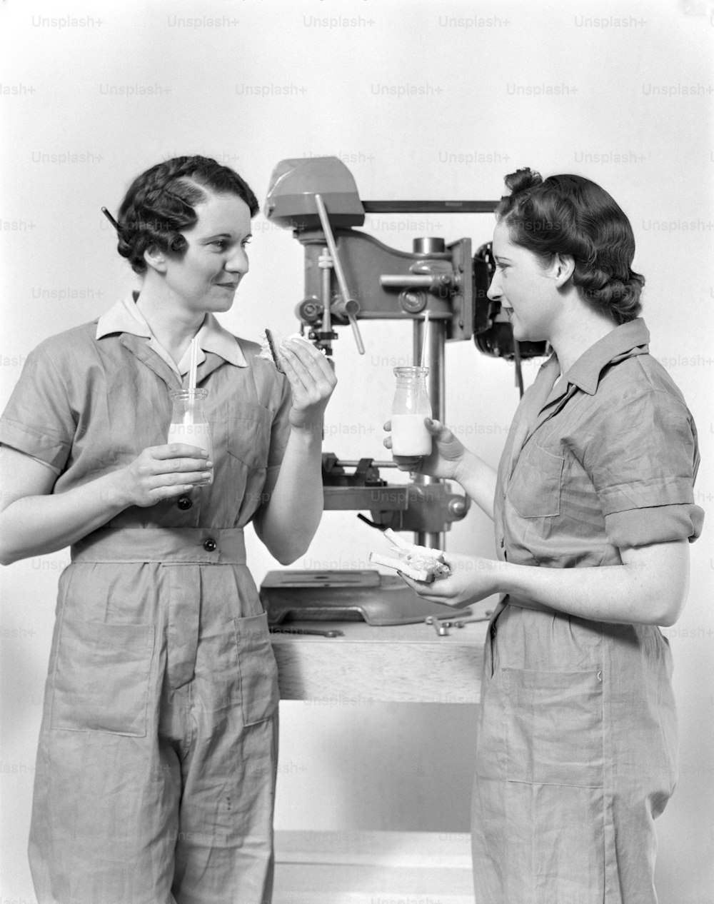 ÉTATS-UNIS - Vers les années 1940 : Deux ouvrières debout près d’une perceuse à colonne, mangeant un sandwich et buvant du lait.