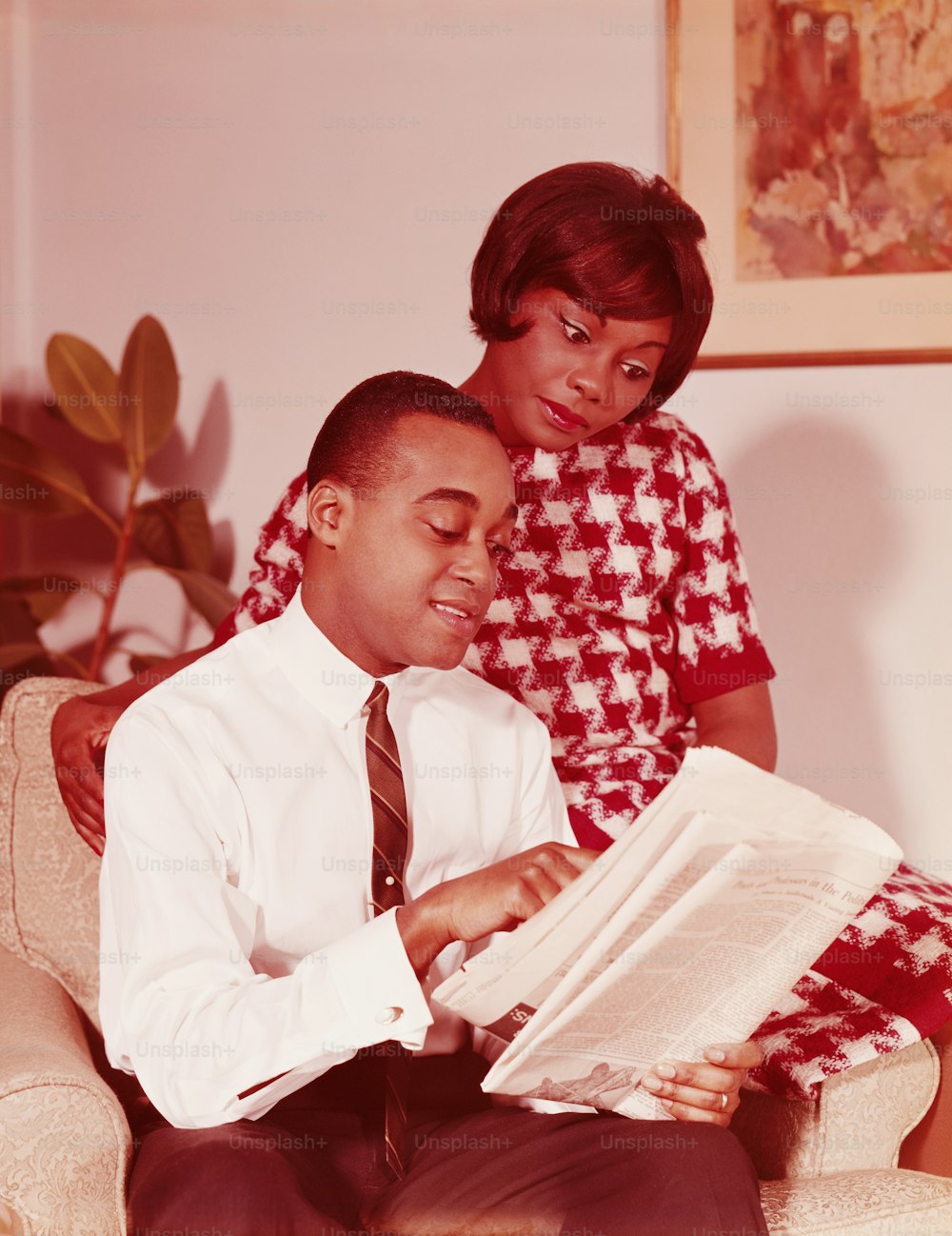 STATI UNITI - CIRCA 1960: Coppia seduta sulla sedia, uomo che legge il giornale, donna sul bracciolo appollaiato della sedia, guardando oltre la spalla dell'uomo.