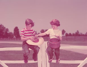 UNITED STATES - CIRCA 1950s:  Boy and girl sitting on farm fence, with basket of cherries.