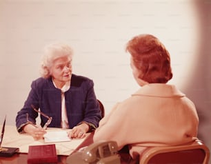 UNITED STATES - CIRCA 1960s:  Two businesswomen seated at desk, talking.