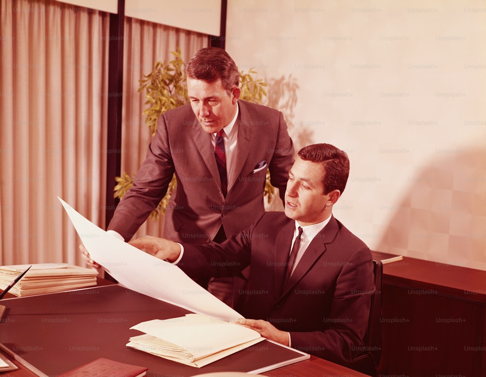 VEREINIGTE STAATEN - CIRCA 1960er Jahre: Zwei männliche Führungskräfte im Büro schauen sich Papiere am Schreibtisch an.