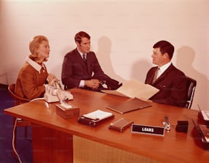 1970年代頃、机に座り、銀行の支店長と話すカップル。