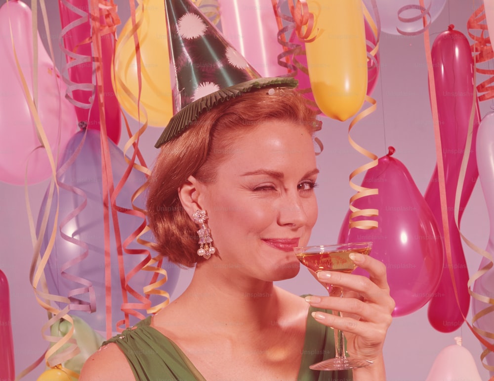 ÉTATS-UNIS - Circa 1960s : Femme à la fête, portant un chapeau de fête et faisant un clin d’œil, tenant un verre de vin.