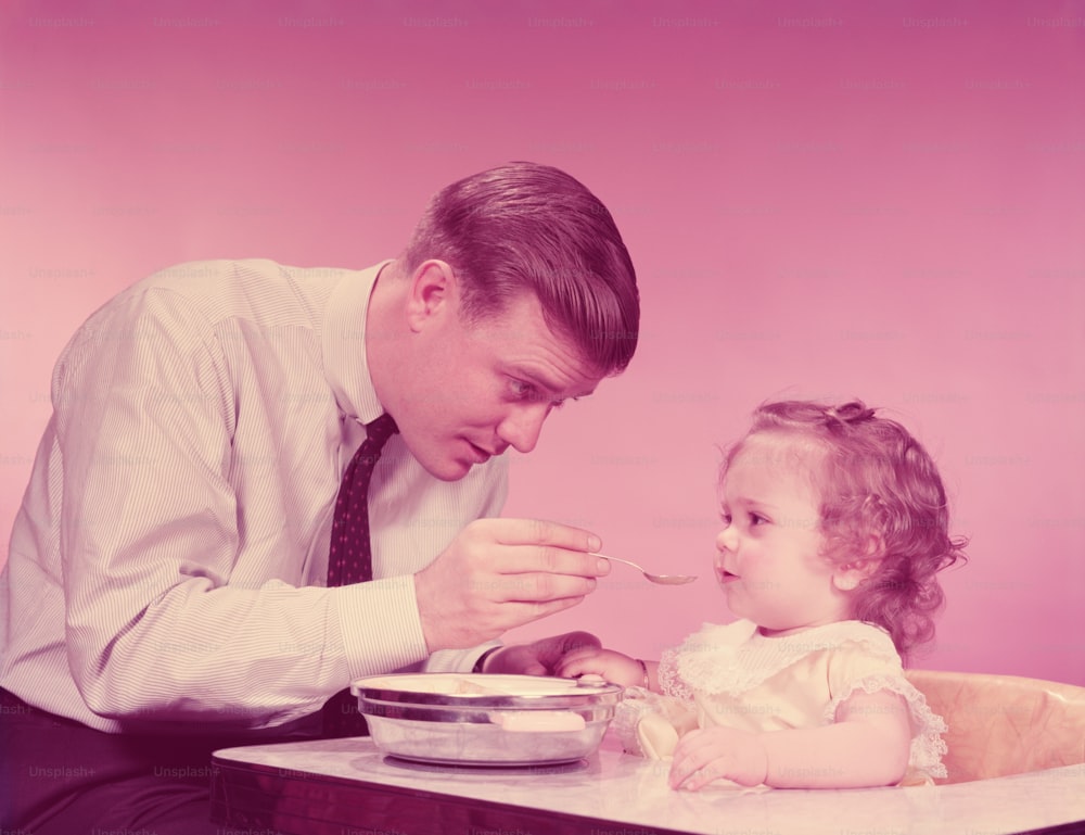 UNITED STATES - CIRCA 1960s:  Father feeding baby girl, sitting in high chair.