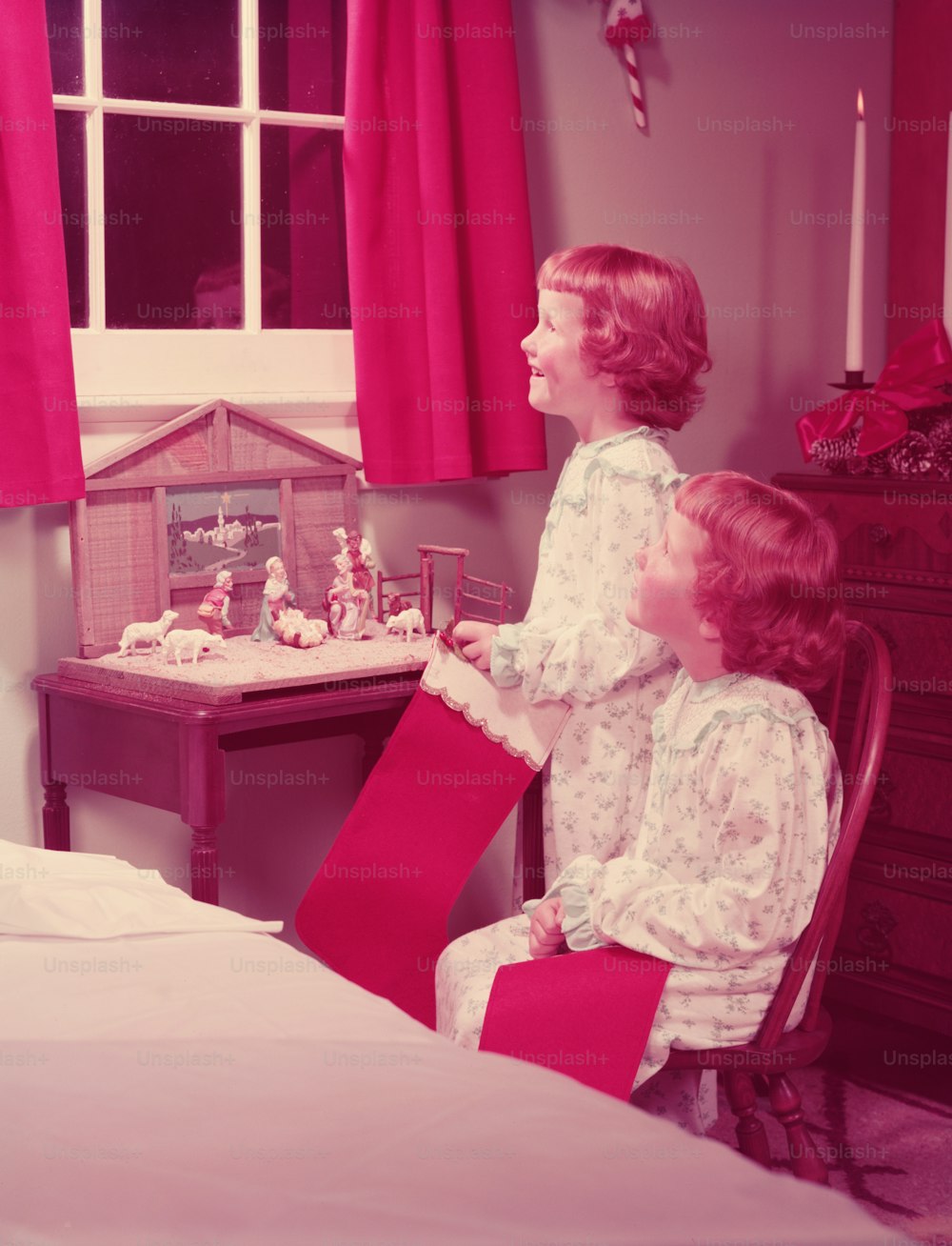 UNITED STATES - CIRCA 1950s:  Twin girls holding Christmas stockings, looking out of bedroom window, nativity scene on night table.
