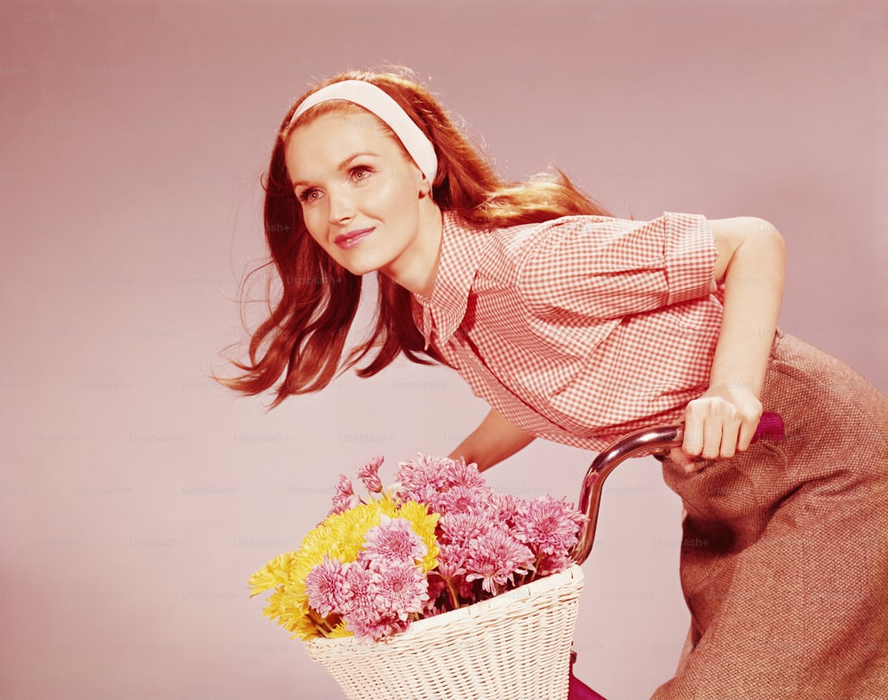 UNITED STATES - CIRCA 1970s:  Young woman on bicycle with basket full of chrysanthemum flowers.