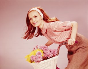 UNITED STATES - CIRCA 1970s:  Young woman on bicycle with basket full of chrysanthemum flowers.