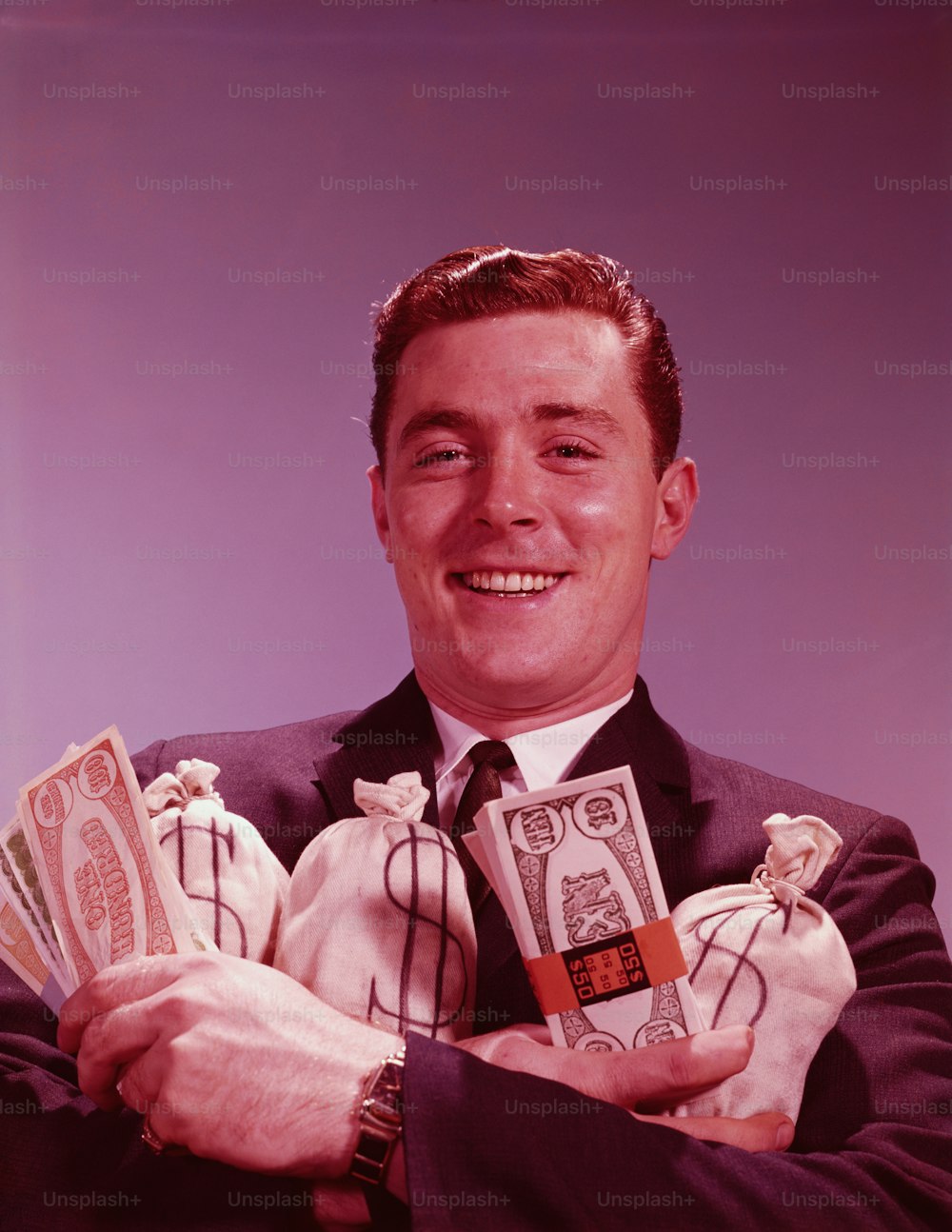 UNITED STATES - CIRCA 1960s:  Man holding bundles and bags of money, smiling, portrait.