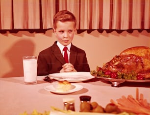 ÉTATS-UNIS - Vers les années 1960 : Garçon assis à table, les mains jointes pour la prière de grâce, regardant une dinde rôtie.