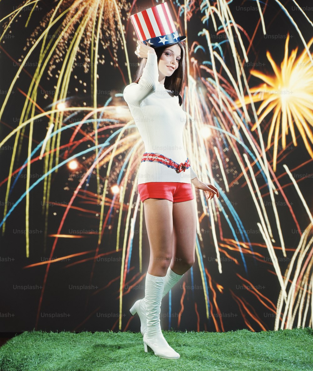 ETATS-UNIS - Vers les années 1970 : Jeune femme en pantalon chaud et bottes go-go, portant un chapeau de l’Oncle Sam, feux d’artifice en arrière-plan.