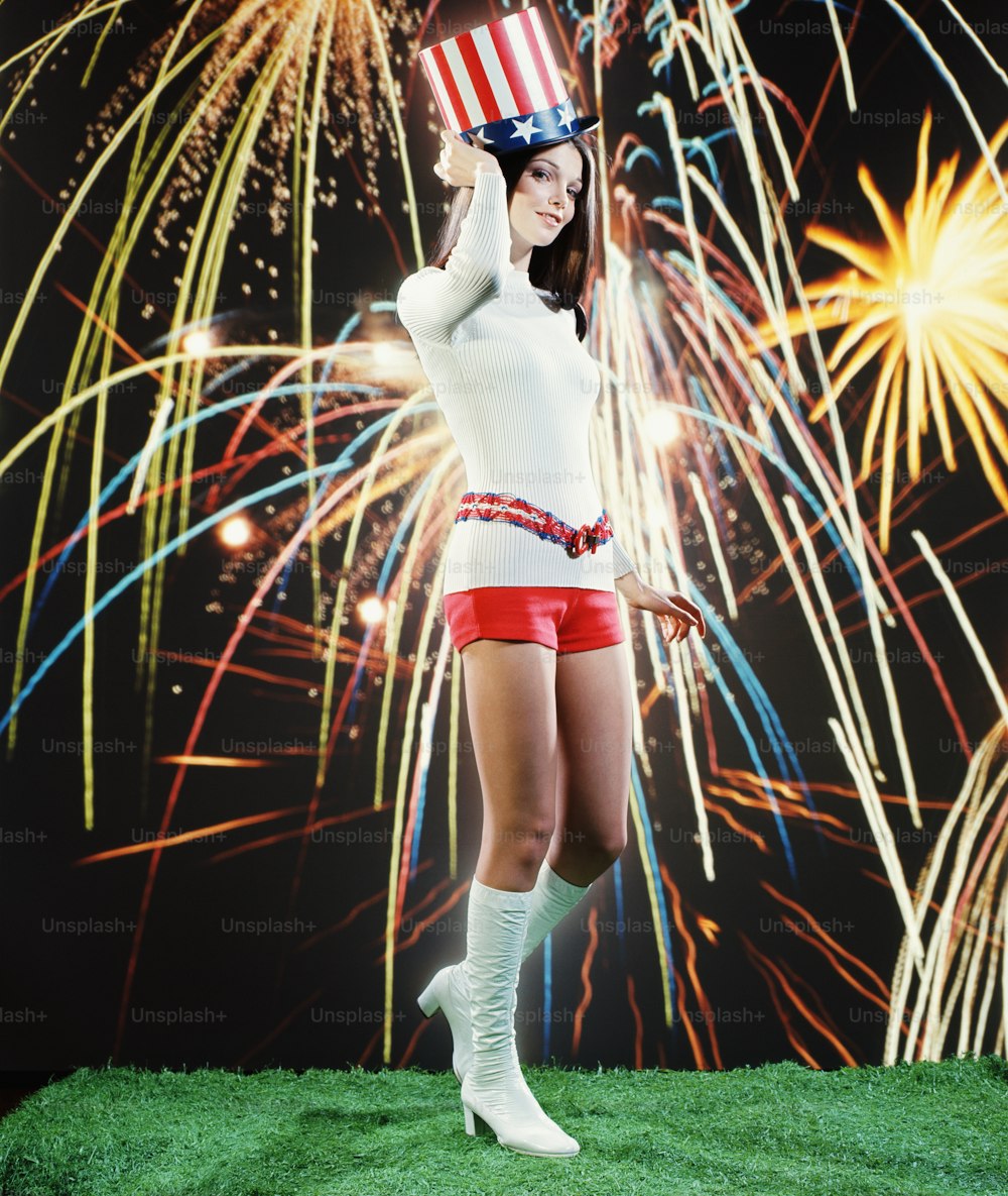 UNITED STATES - CIRCA 1970s:  Young woman in hot pants and go-go boots, wearing Uncle Sam hat, fireworks in background.