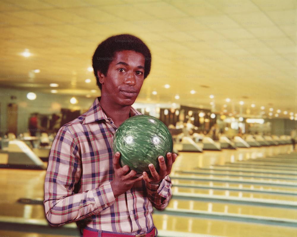 ESTADOS UNIDOS - POR VOLTA DE 1970: Homem segurando bola de boliche verde em mármore na pista de boliche, pistas ao fundo.
