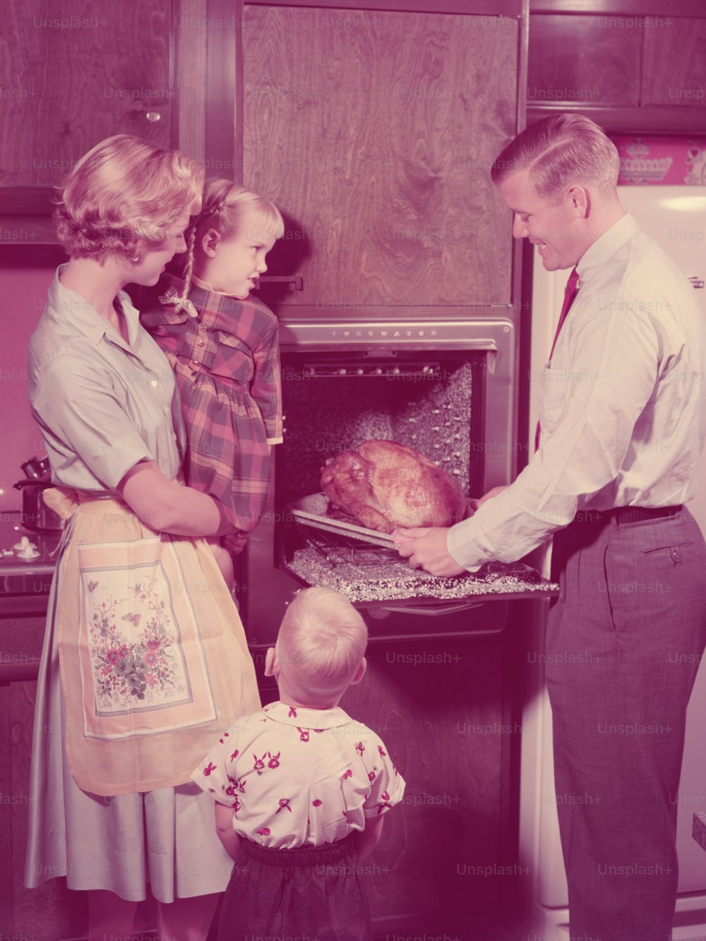 UNITED STATES - CIRCA 1950s:  Family in kitchen, father taking roast turkey out of oven, mother and children looking on.