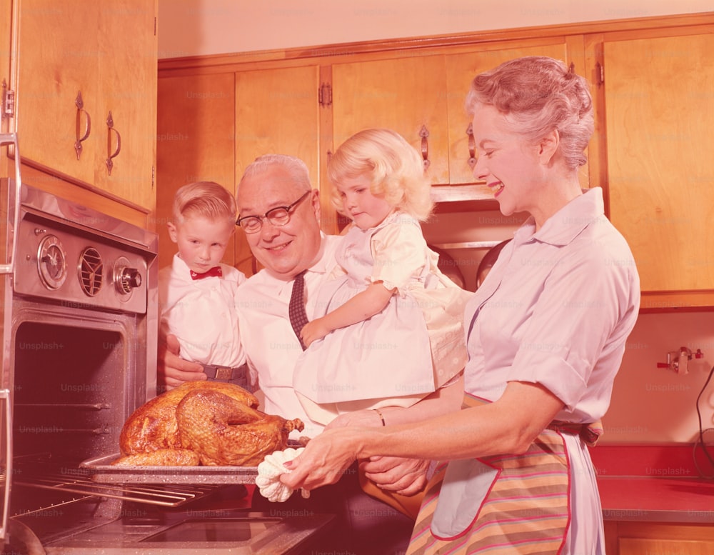 UNITED STATES - CIRCA 1950s:  Grandfather holding grandchildren and looking, grandmother removing roast turkey from oven.