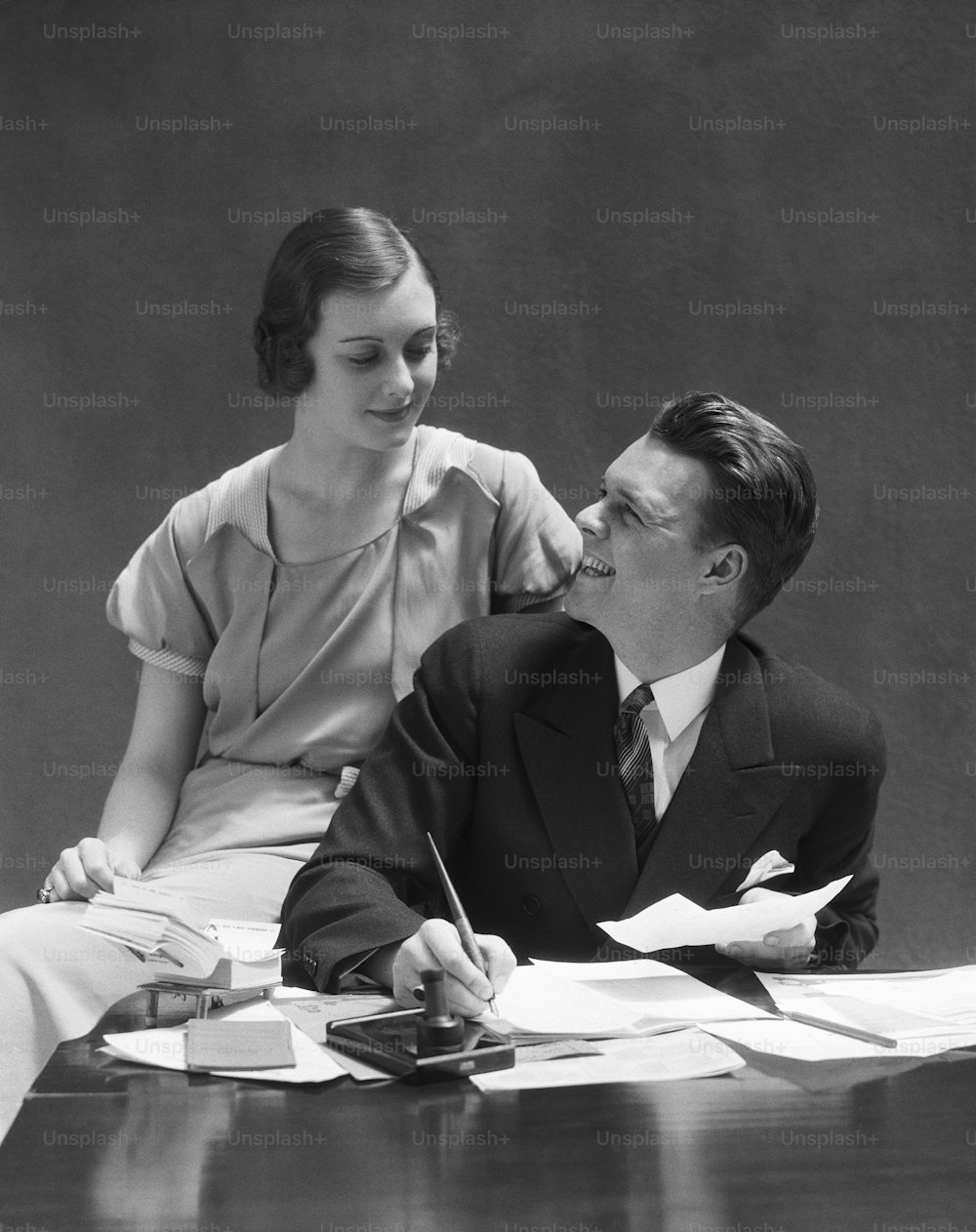 미국 - 1930년대경: 책상에 앉아 있는 커플, 의자 팔에 앉은 여자를 올려다보는 남자.