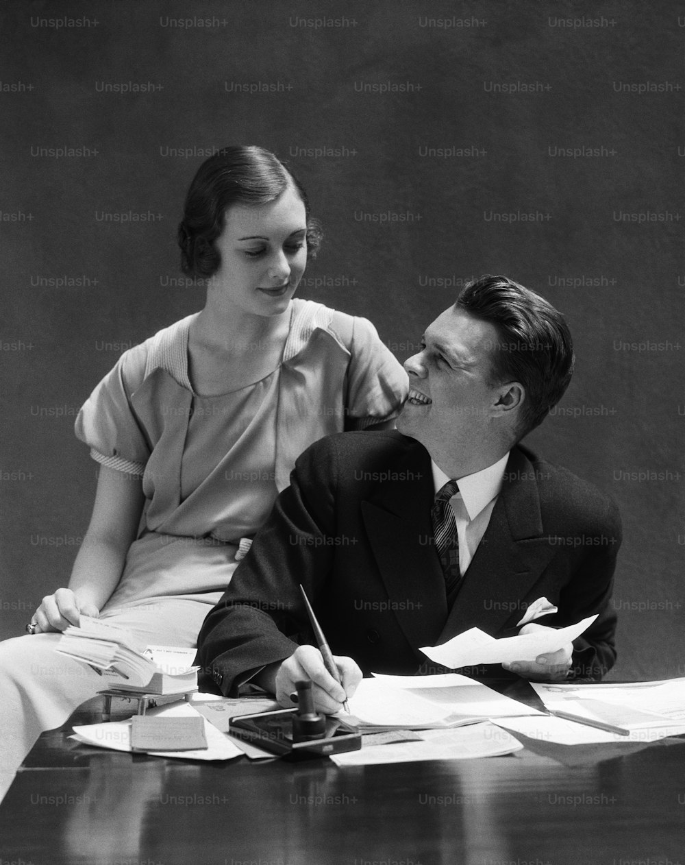 UNITED STATES - CIRCA 1930s:  Couple sitting at desk, man looking up at woman seated on arm of the chair.