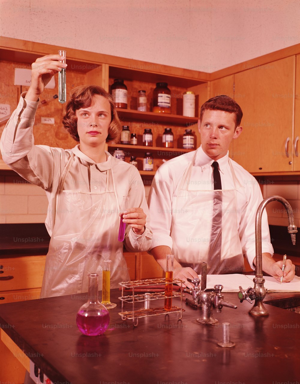 UNITED STATES - CIRCA 1960s:  Two students in chemistry laboratory, girl holding up test tube, boy taking notes science.