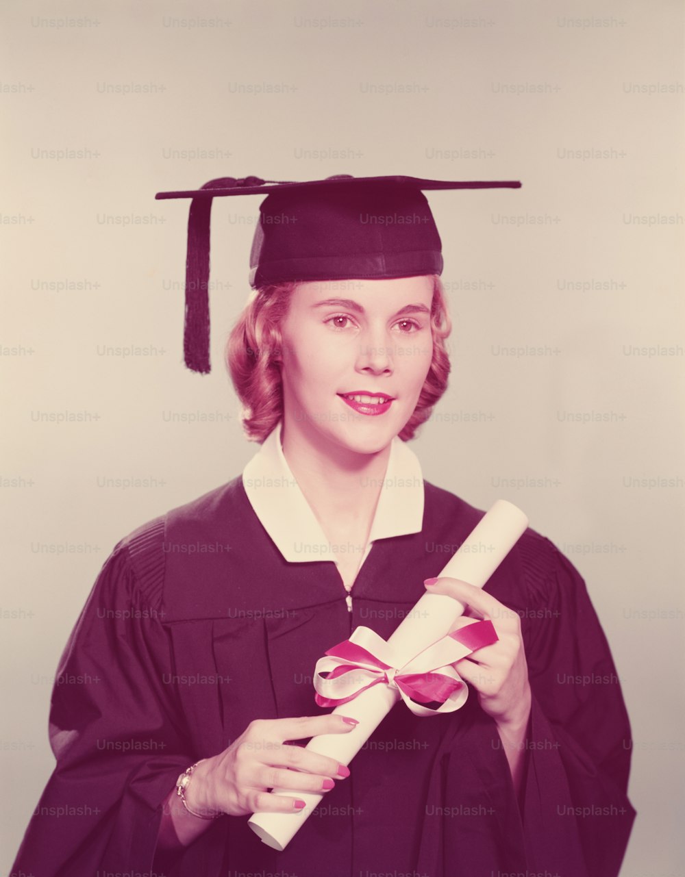 UNITED STATES - CIRCA 1950s:  Young woman wearing graduation robes and mortarboard, holding diploma tied with red and white ribbon.