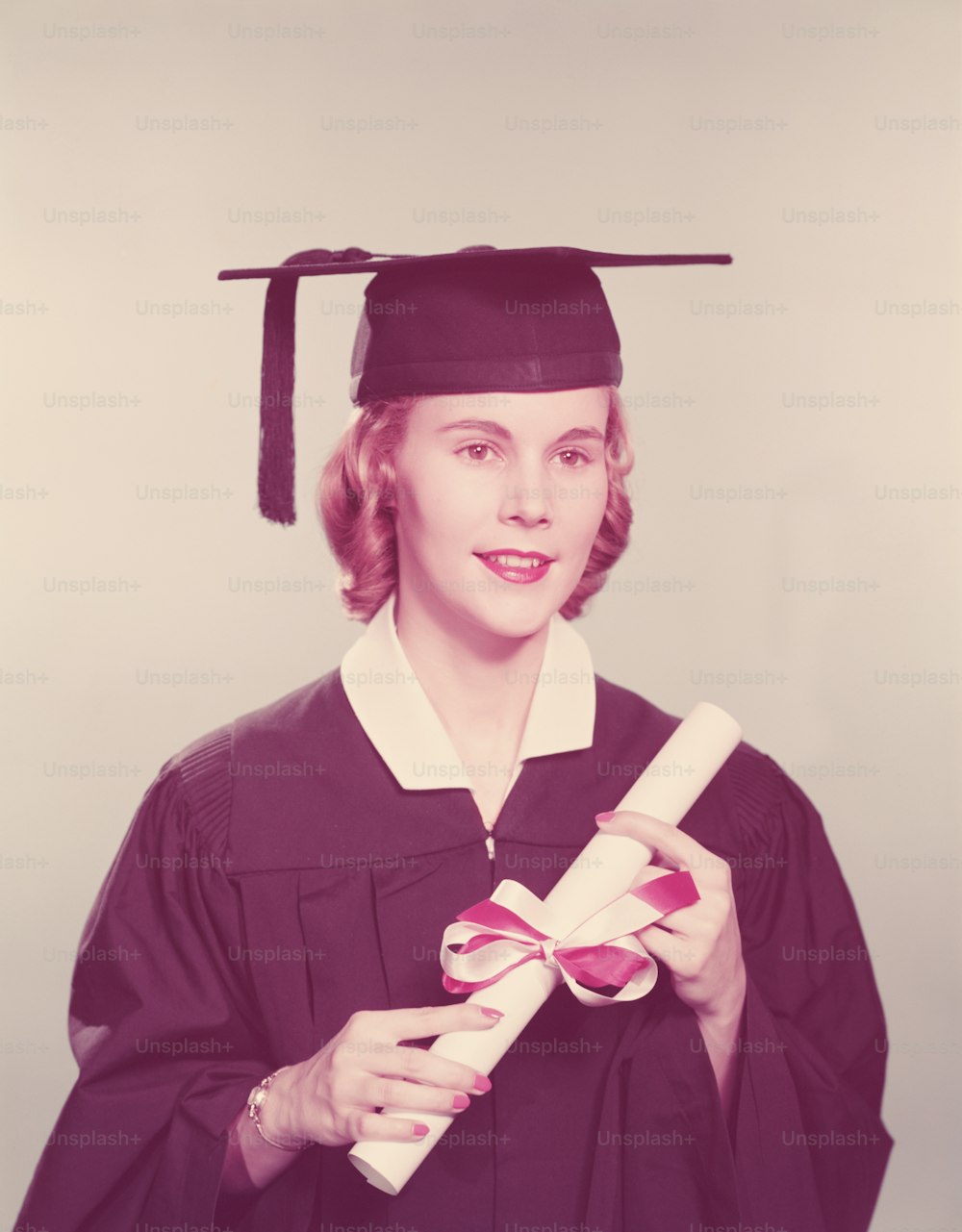UNITED STATES - CIRCA 1950s:  Young woman wearing graduation robes and mortarboard, holding diploma tied with red and white ribbon.