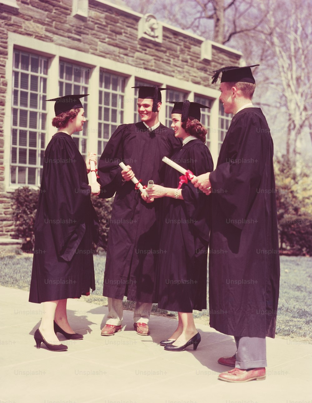UNITED STATES - CIRCA 1950s:  Four students wearing graduation robes and mortarboards, holding diplomas outside school building.